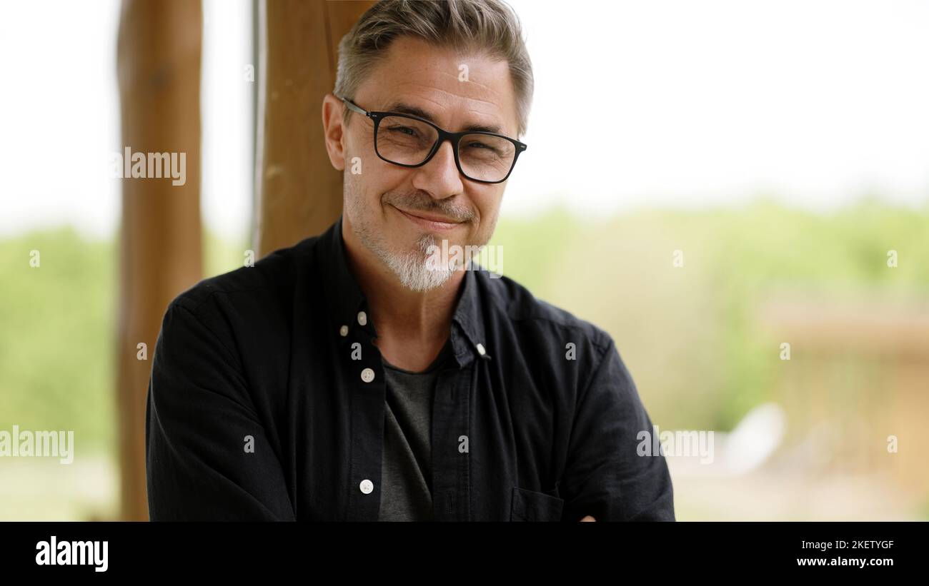 Portrait of happy casual middle age man in glasses standing on balcony at home relaxing, smiling. Stock Photo