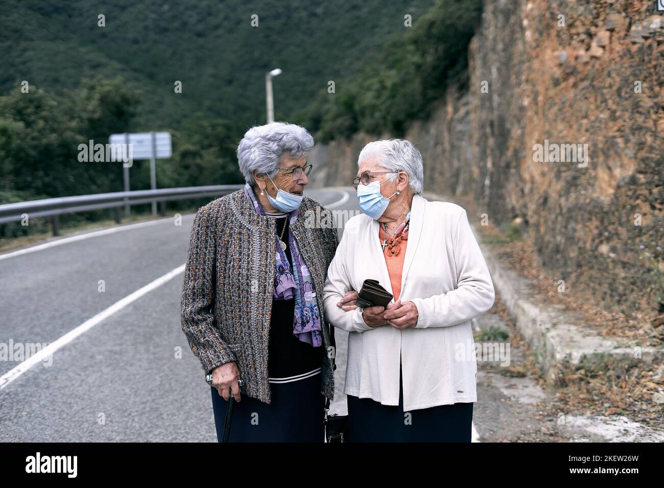 two caucasian elderly ladies with white hair and glasses talking calmly next to the dangerous road that passes through the forest with face mask, ruta Stock Photo