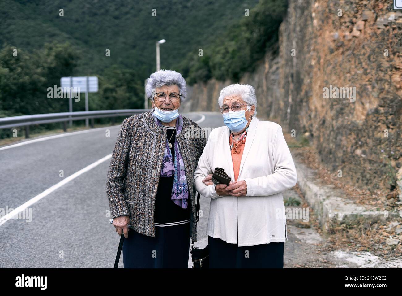 two elderly caucasian ladies with white hair and glasses walking on the dangerous road next to the forest smiling looking at camera with face mask Stock Photo