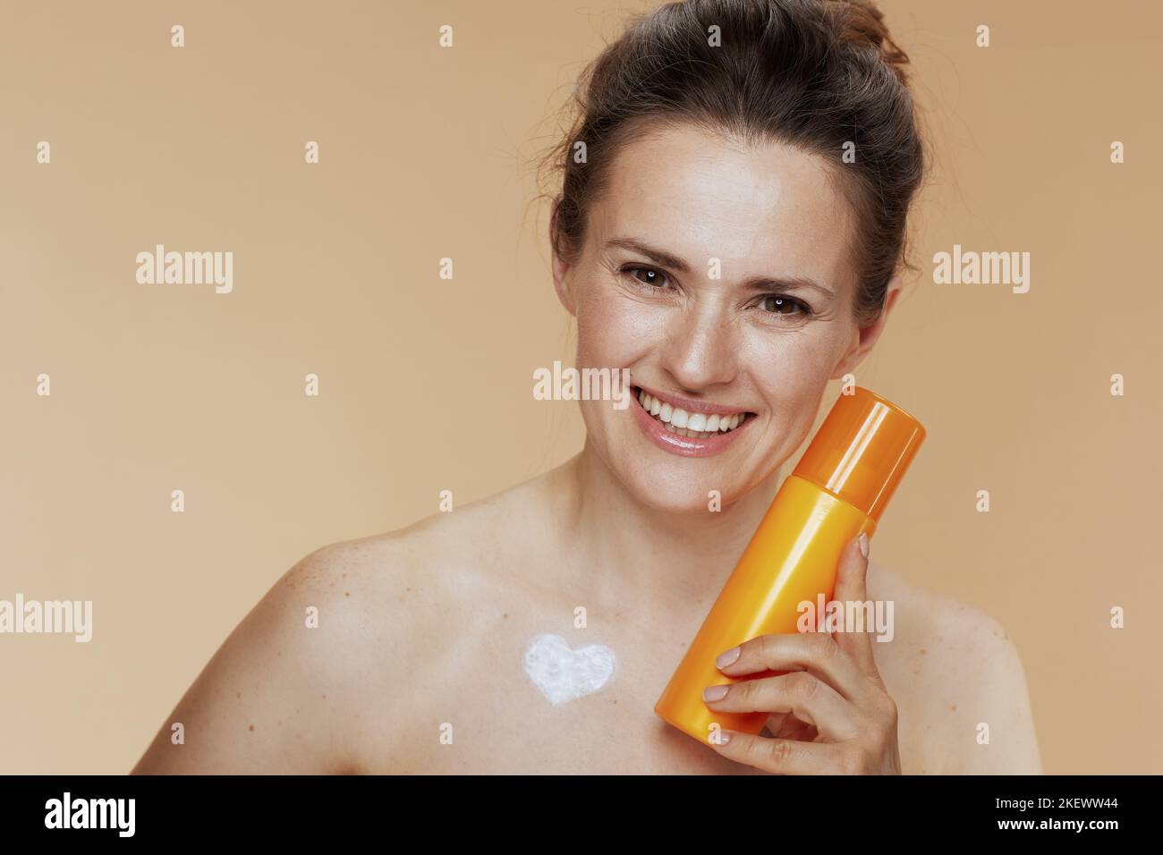 happy modern woman with cosmetic cream jar and heart shaped cream on chest on beige background. Stock Photo