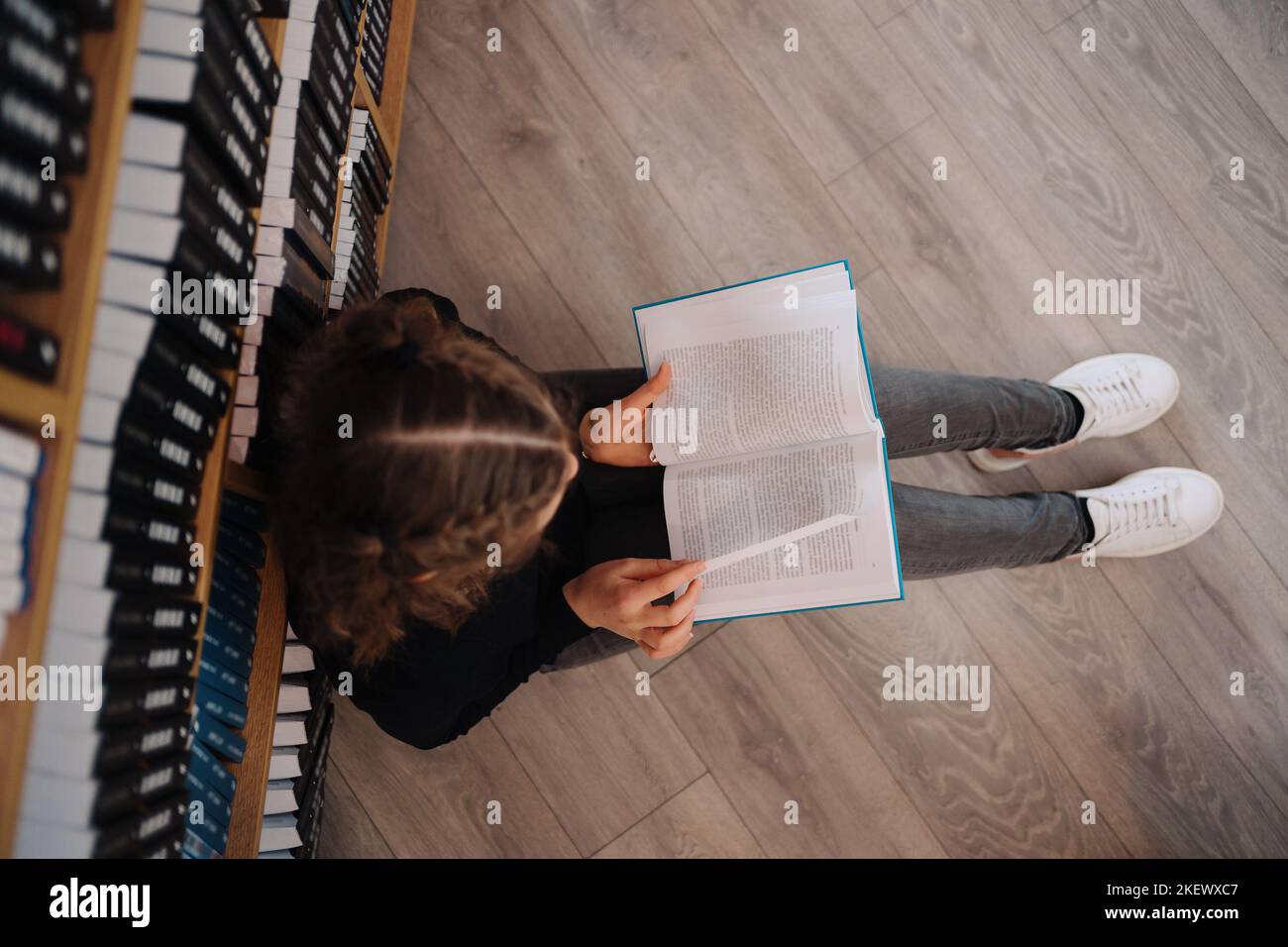 Teen girl among a pile of books. A young girl wearing glasses reads a book with shelves in the background. She is surrounded by stacks of books. Book Stock Photo