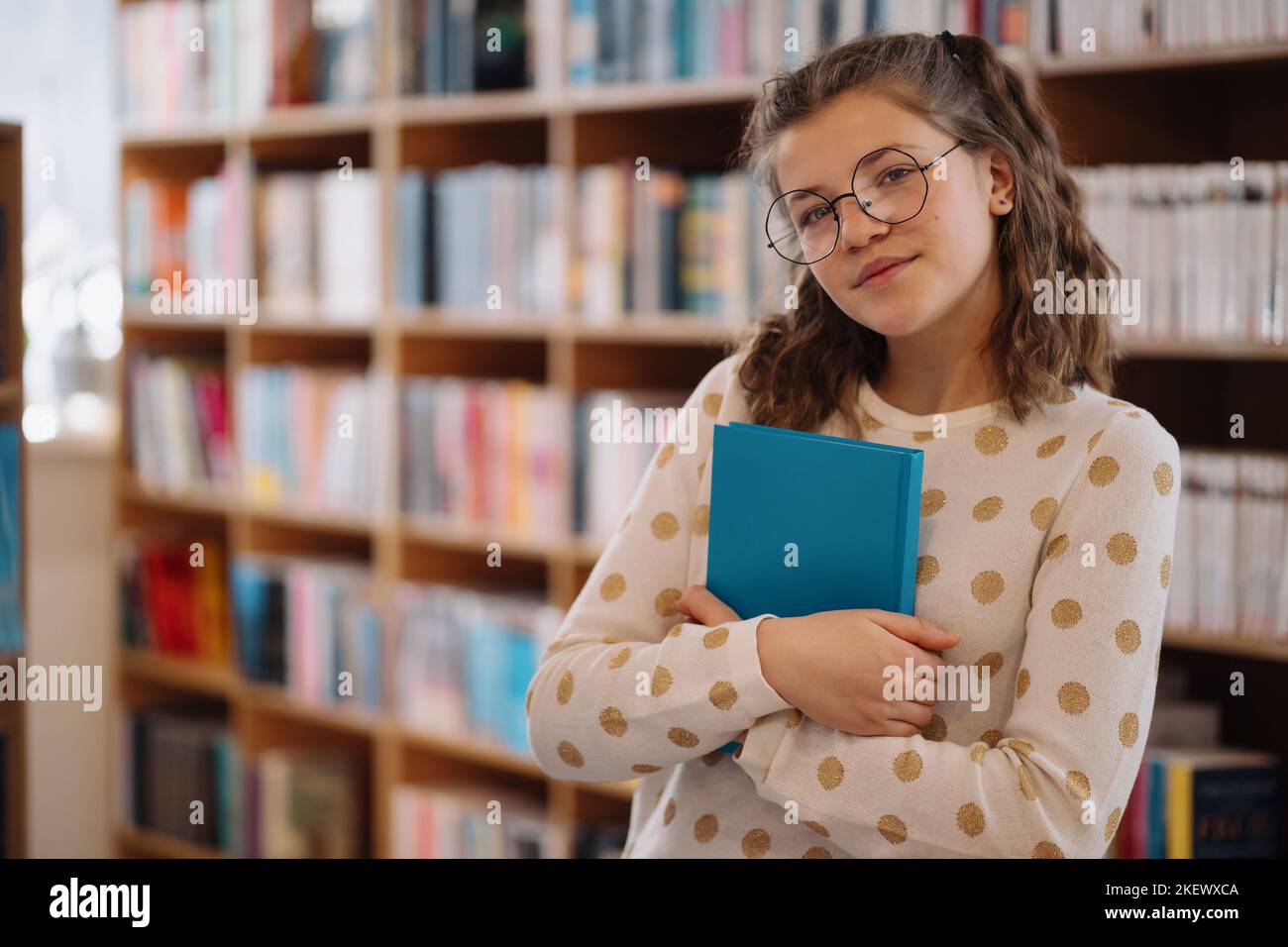 Teen girl among a pile of books. A young girl holding book with shelves in the background. She is surrounded by stacks of books. Book day. Stock Photo