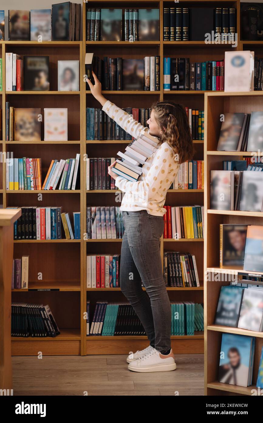 Teen girl among a pile of books. A young girl holding books with shelves in the background. She is surrounded by stacks of books. Book day. Stock Photo
