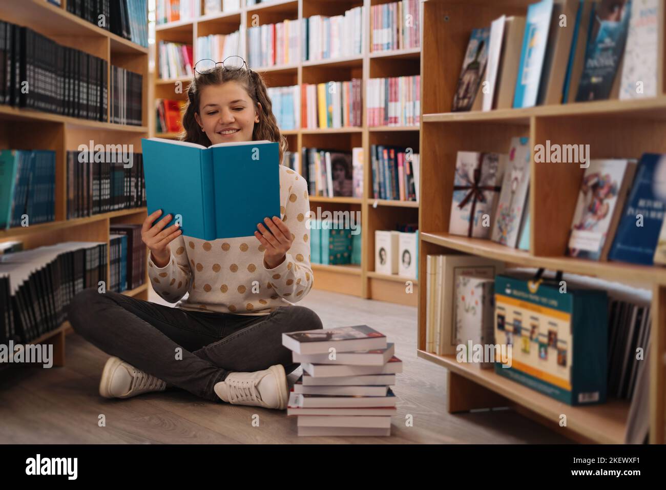 Teen girl among a pile of books. A young girl reads a book with shelves in the background. She is surrounded by stacks of books. Book day. Stock Photo