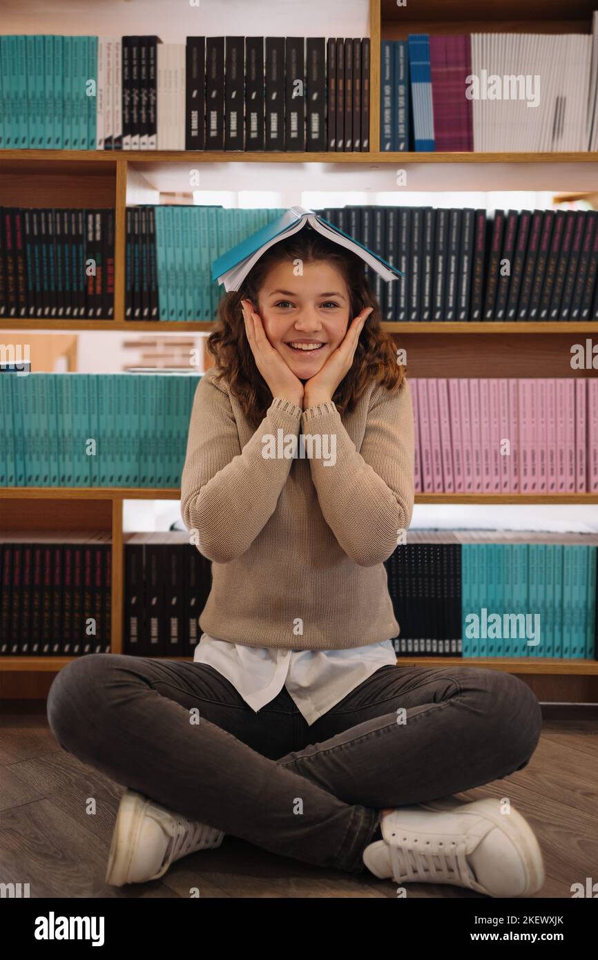 Portrait of a pretty young girl hiding behind an open blue book and looking away over bookshelf background Stock Photo