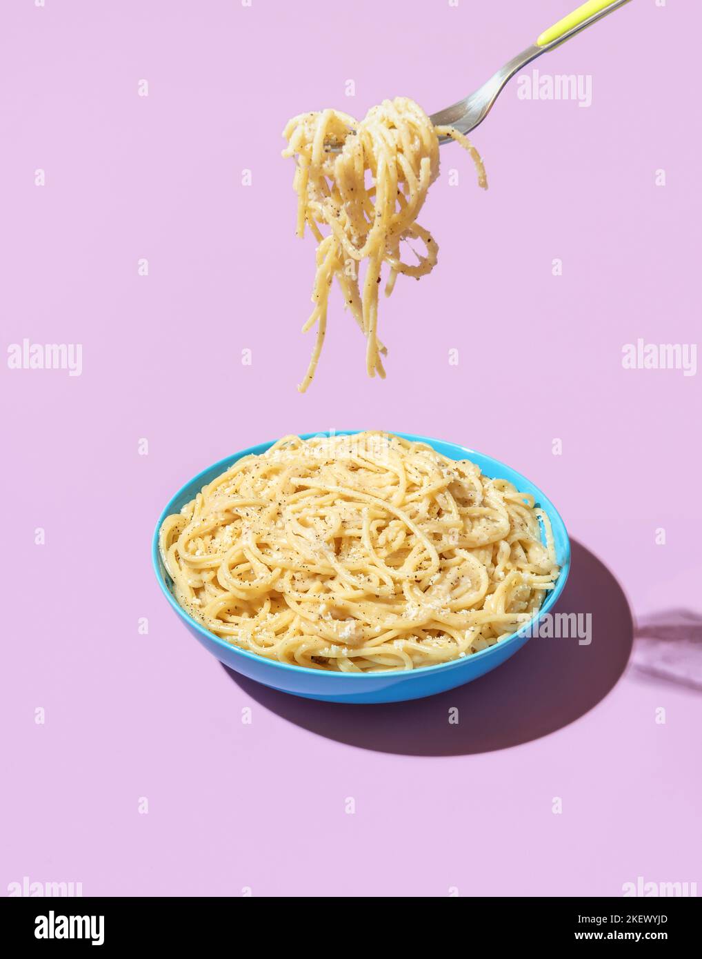 Pasta Cacio e Pepe in a blue bowl isolated on a purple background. Taking spaghetti with a fork from a bowl in bright light against a colorful backgro Stock Photo
