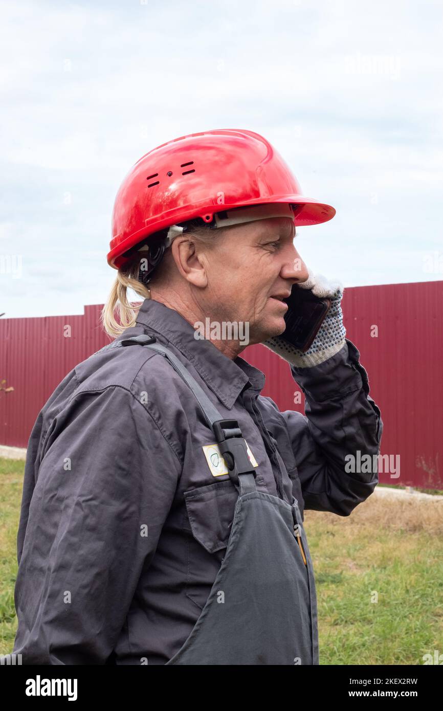 An adult male worker in overalls and a hard hat is talking on the phone on the street. Stock Photo