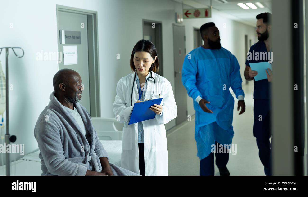 Asian female doctor and smiling senior african american male patient talking in hospital corridor. Hospital, medical and healthcare services. Stock Photo
