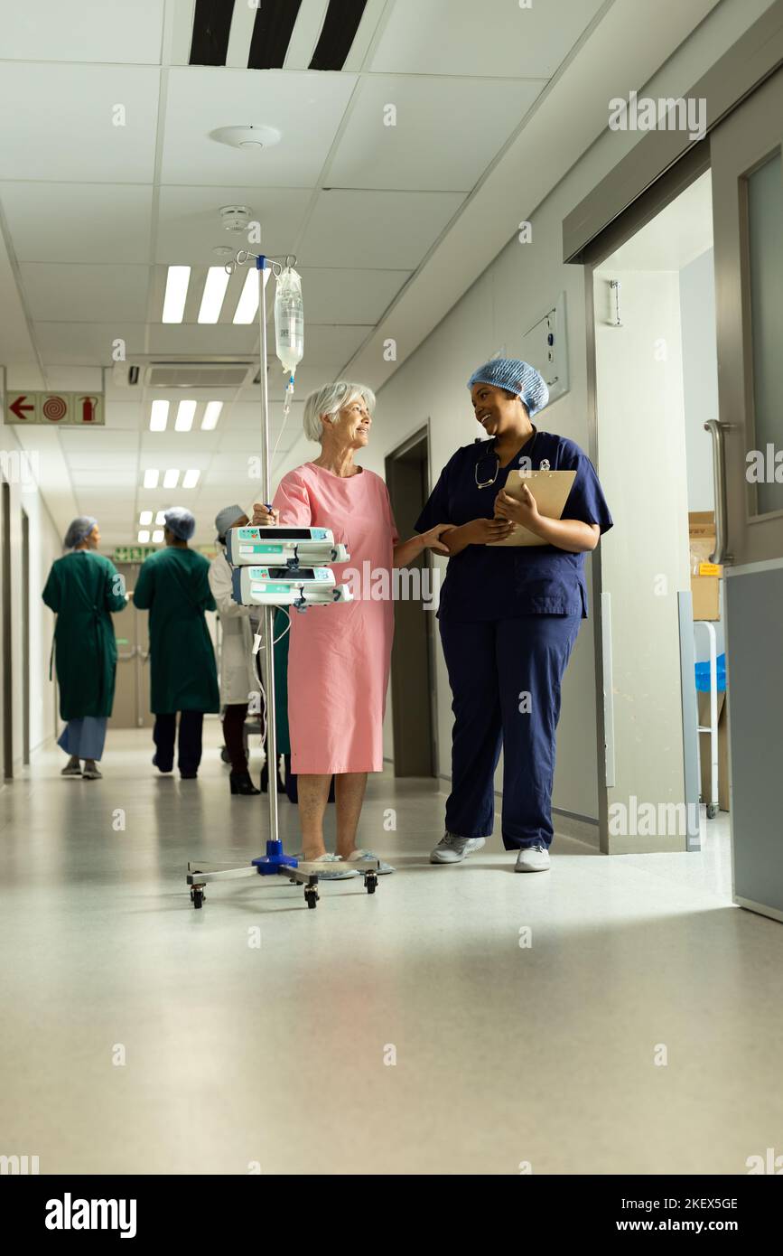 Diverse female doctor and caucasian senior female patient with iv pole talking. Hospital, medical and healthcare services. Stock Photo