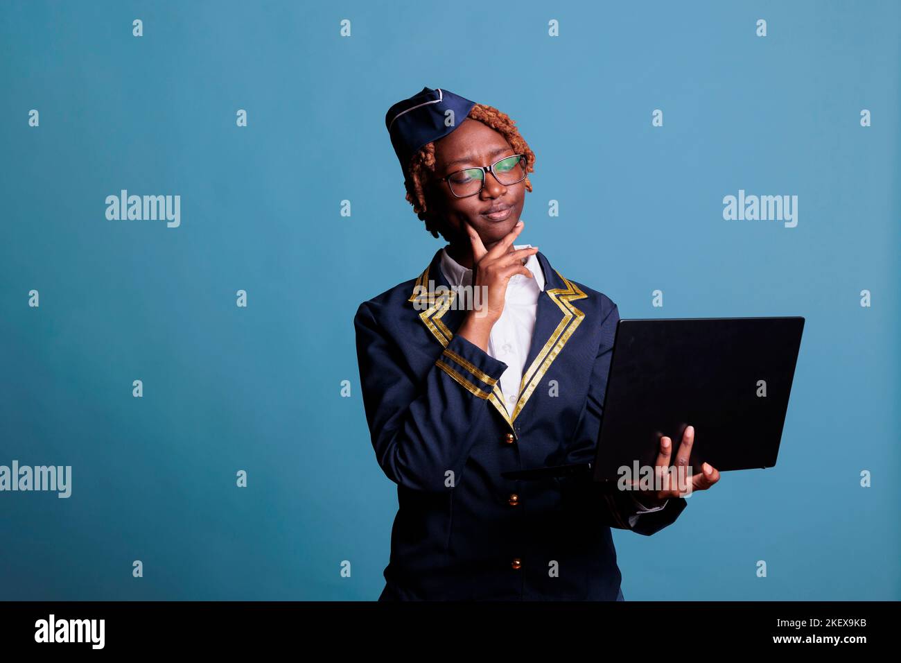 Thoughtful female african american flight attendant in uniform using laptop in studio shot. Stewardess looking doubtfully at new directives received by mail against a blue background. Stock Photo