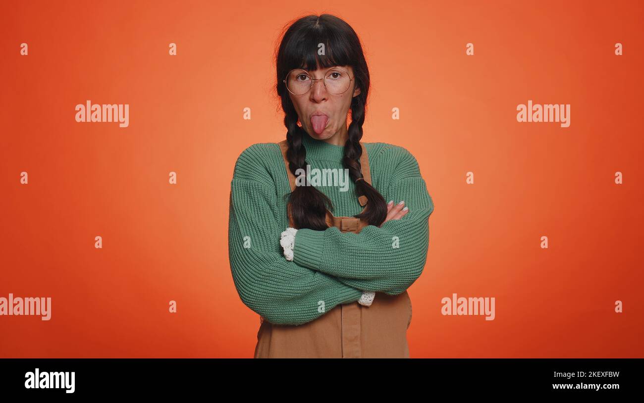 Cheerful funny woman in green sweater showing tongue making faces at camera, fooling around, joking, aping with silly face teasing. Young nerd adult girl isolated alone on orange studio background Stock Photo