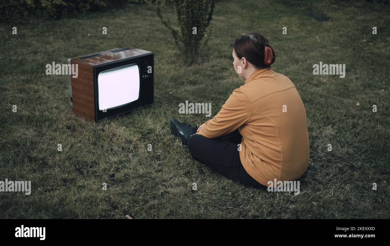 Woman is sitting on the grass and watching an old retro TV. Concept of horror Stock Photo