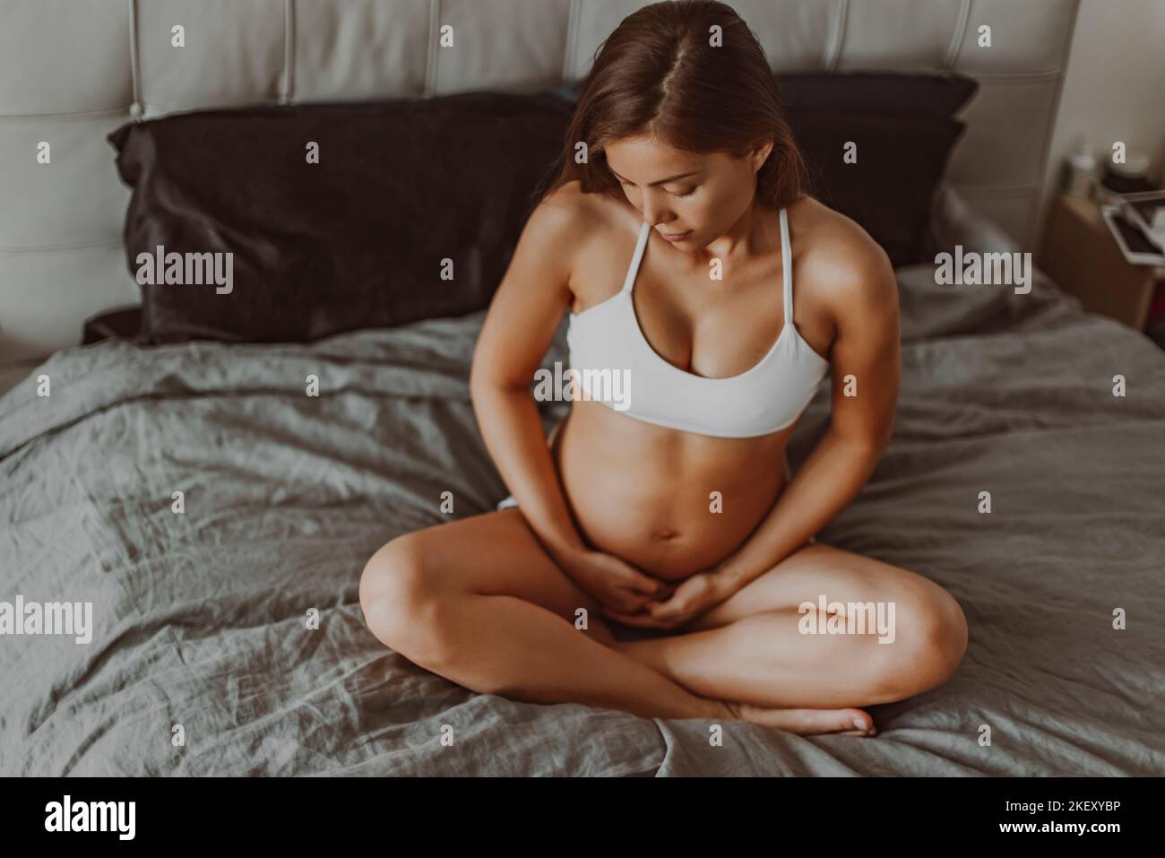 Pregnant woman sitting in bed during pregnancy thinking about her baby and motherhood. Happy multiracial asian model in first trimester showing belly Stock Photo