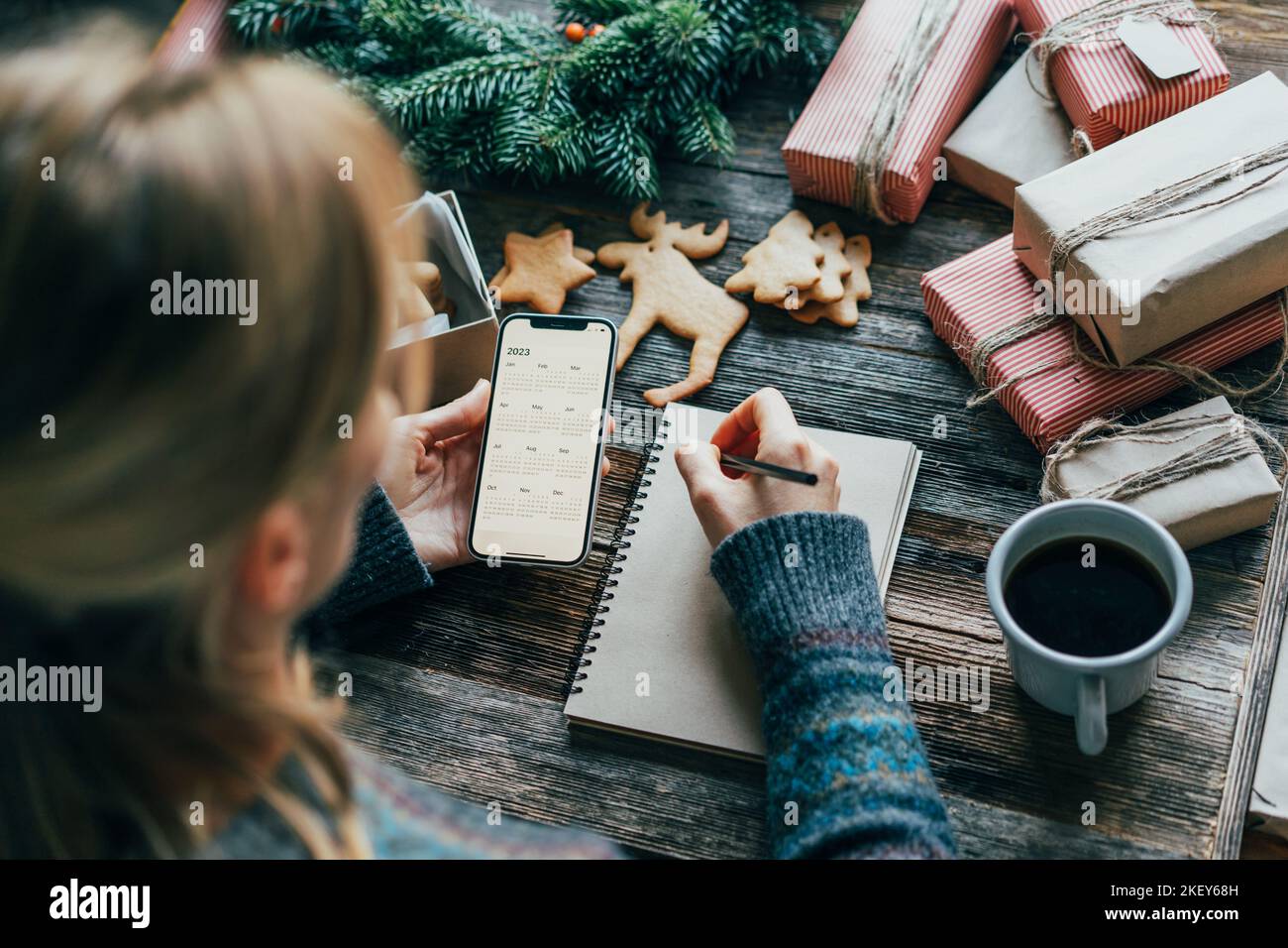 Woman writing plans for 2023 on the Christmas table, holding a phone with a calendar on the screen. Stock Photo