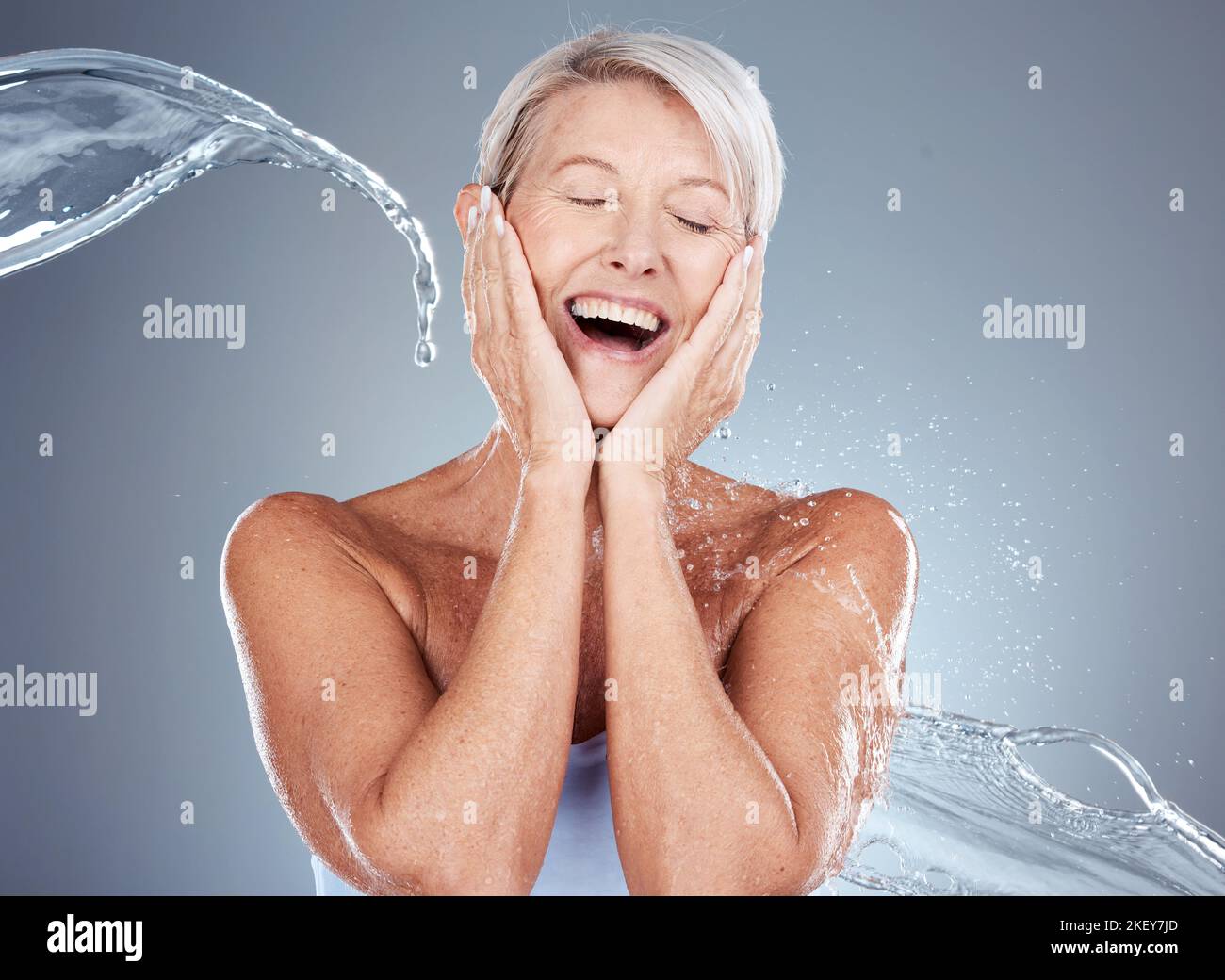 Mature woman, water and splash with shower for clean aesthetic health on a grey background. Beauty, bath and hygiene with a woman washing for bodycare Stock Photo