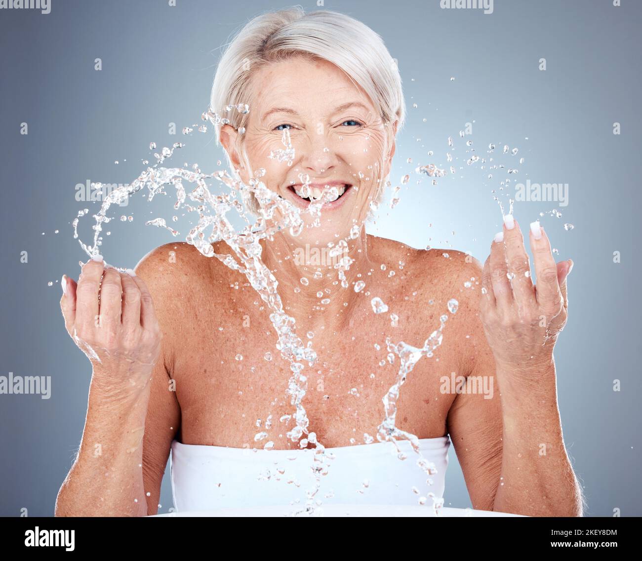 Mature woman, water and splash for beauty, skincare and hygiene or grooming on a grey studio background. Senior woman, fresh and facial wash or Stock Photo