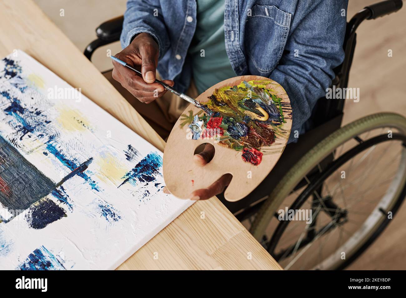 Above view of young black man with color palette mixing acrylic paints while sitting on wheelchair by wooden table with canvas Stock Photo