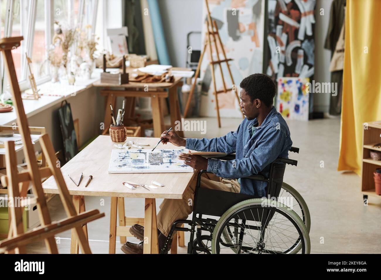 Young contemporary artist in wheelchair working over new painting while sitting in studio of arts, mixing paints and applying them on canvas Stock Photo