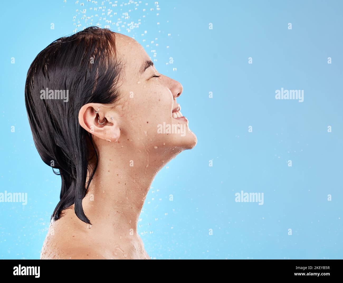 Asian woman, shower and water for cleaning, smile and happy in blue studio mockup background. Model, clean and showering face, body and hygiene Stock Photo