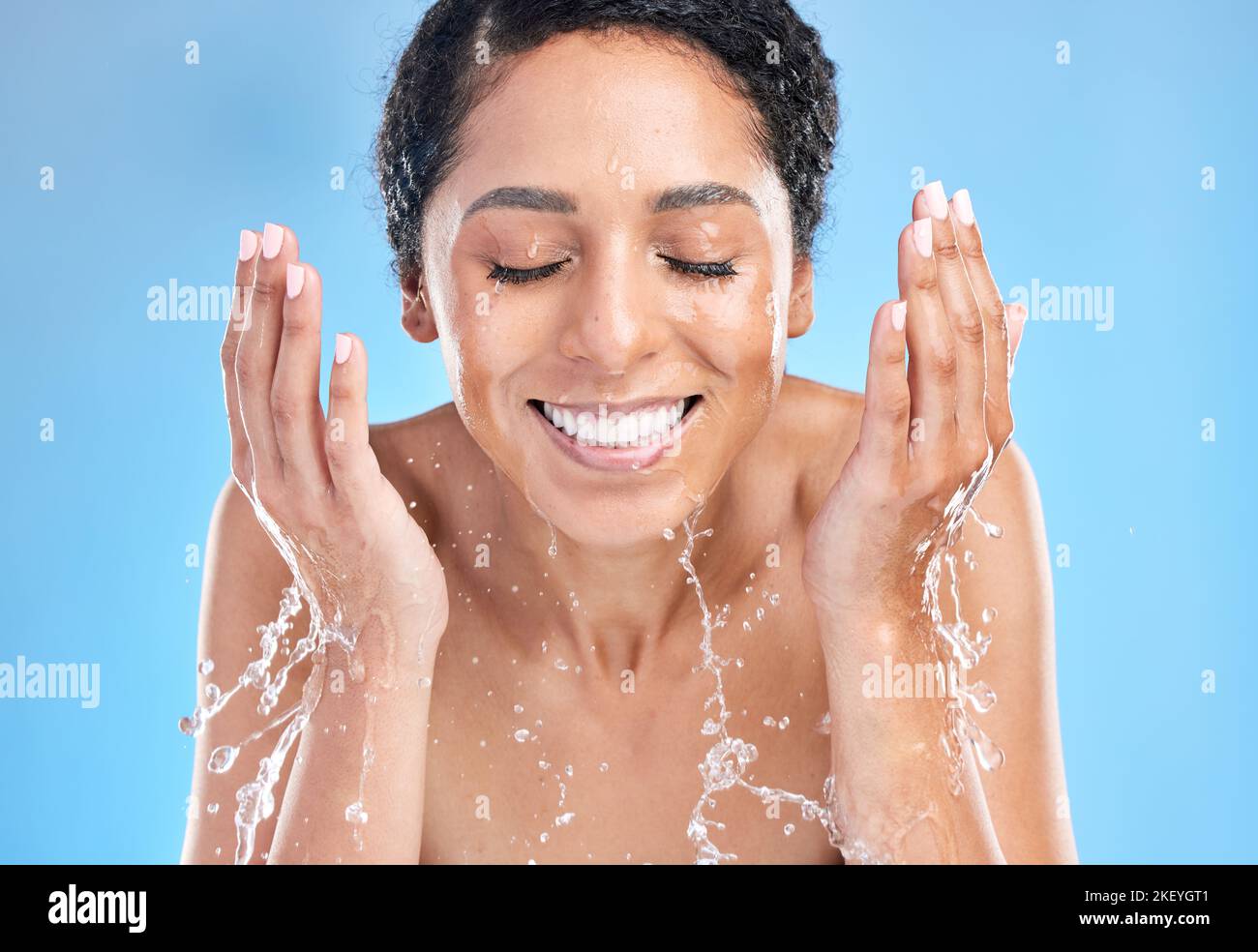 Water splash black woman cleaning face on blue background for health, beauty and skin care morning routine. Skincare, luxury cosmetics and happy woman Stock Photo