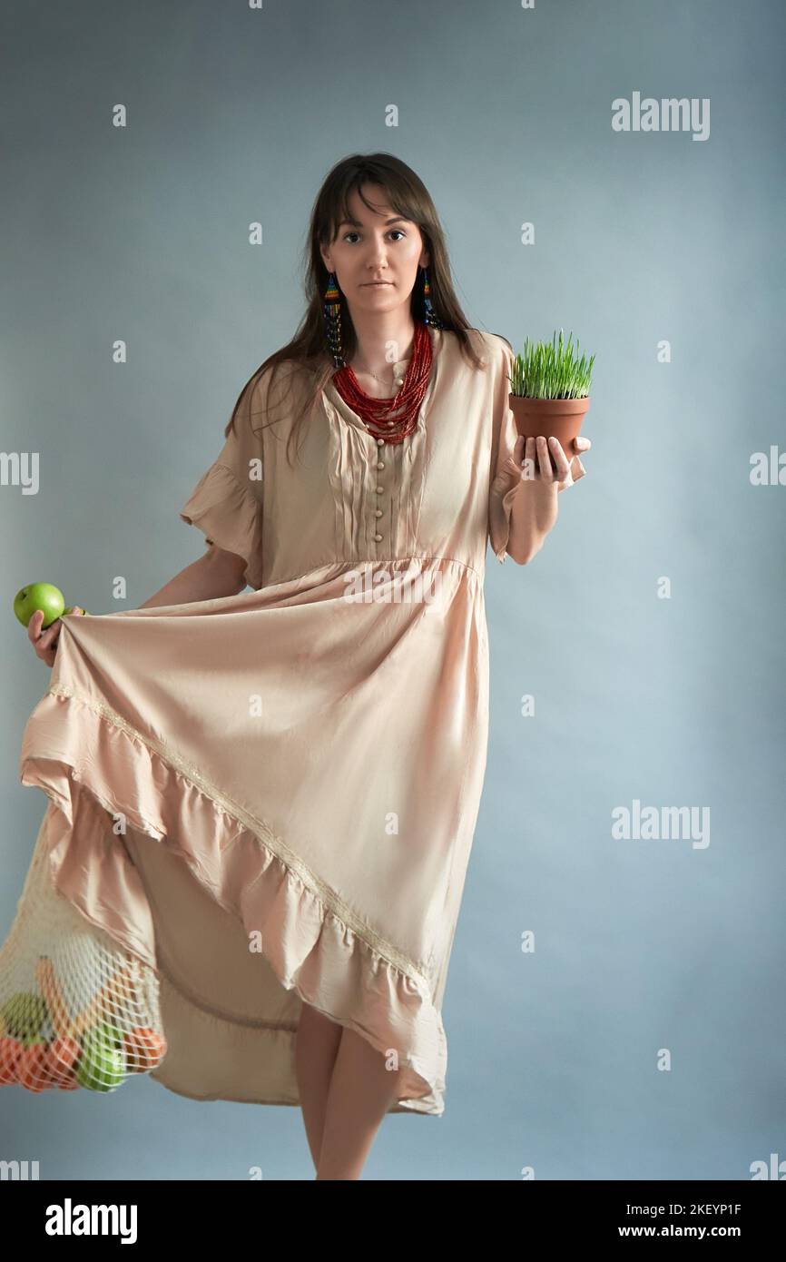 A young long-haired brunette girl in a dress on a gray background with a string bag of fruits and fresh herbs. Shopping for fresh food in a natural products store, advertising ecological cosmetics or a banner for your design. Stock Photo