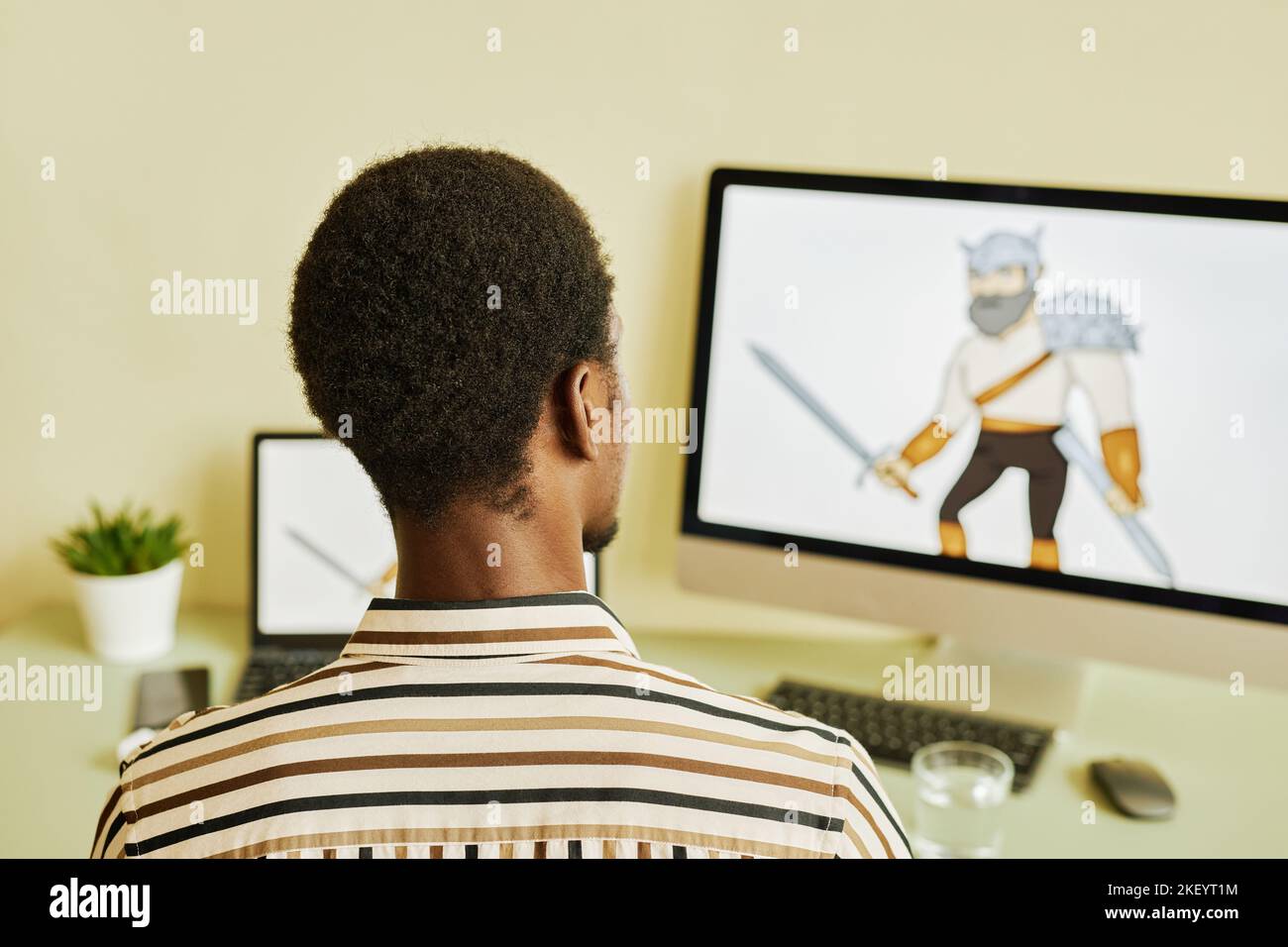 Rear view of young African American designer drawing graphic picture of warrior while sitting in front of computer screen by workplace Stock Photo