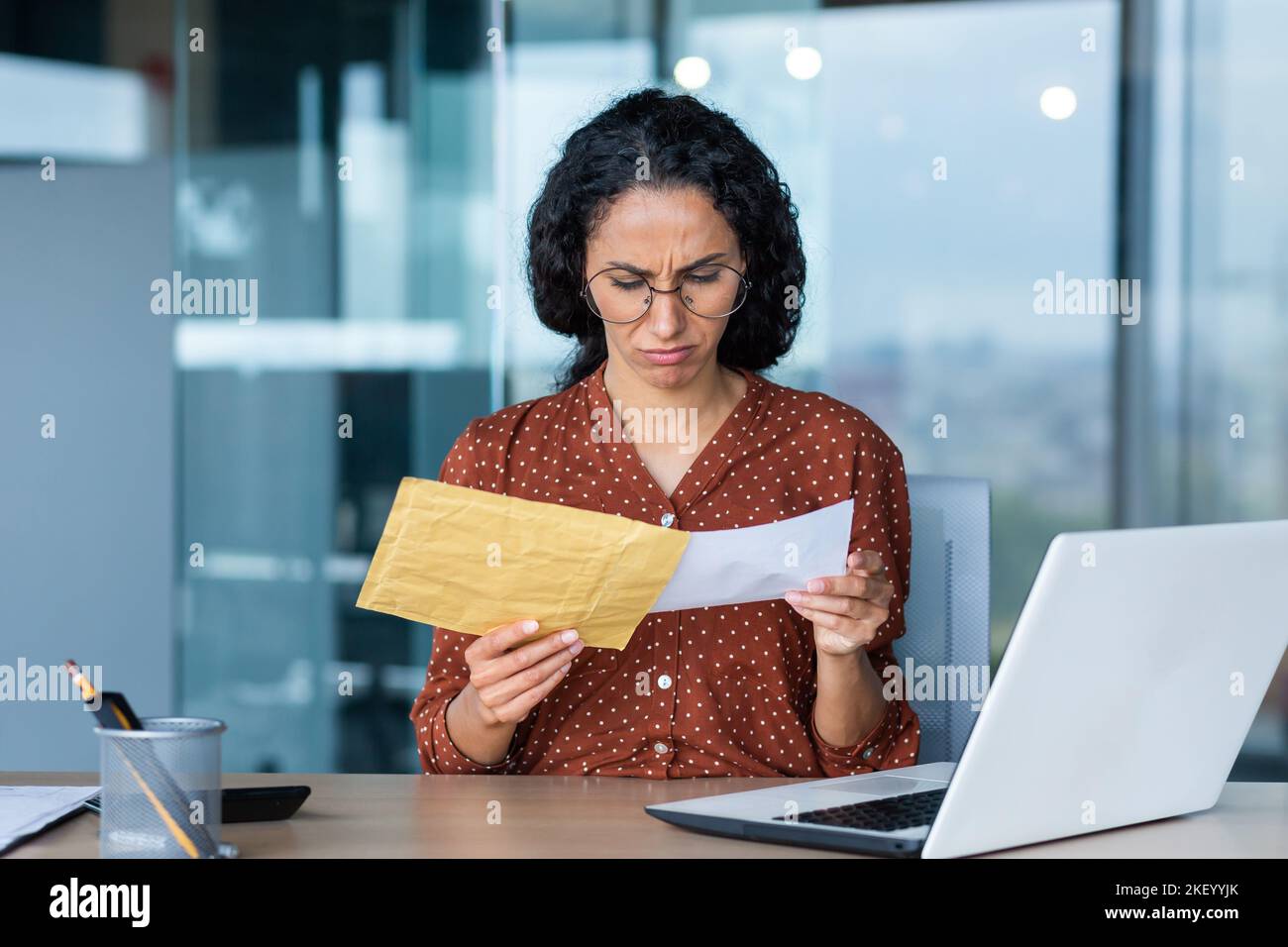 Dissatisfied and upset businesswoman received letter in envelope, depressed latin american businesswoman reading notification message, at work inside office with laptop. Stock Photo