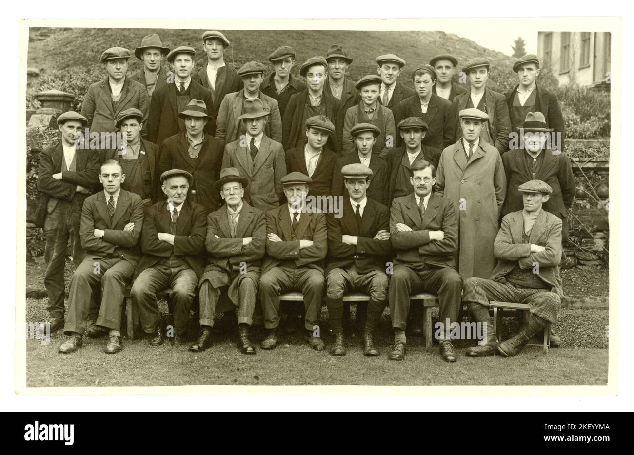 Original and clear 1920's era postcard of a group of estate workers made up of working class men wearing suits, flat caps and boots, and young apprentices/ beaters, and smarter dressed managers, office staff, gamekeeper, (wearing thick woollen socks). Moorland behind and stone balustrade, so maybe estate workers. Lots of characters. U.K Stock Photo