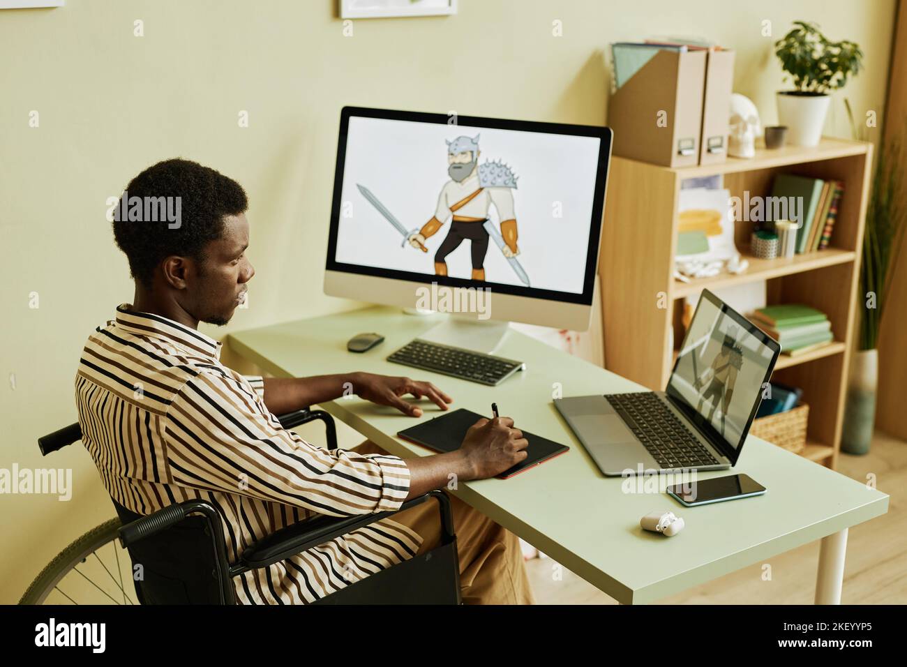 Young serious digital artist with disability looking at laptop screen with graphic picture of warrior while sitting in wheelchair by workplace Stock Photo