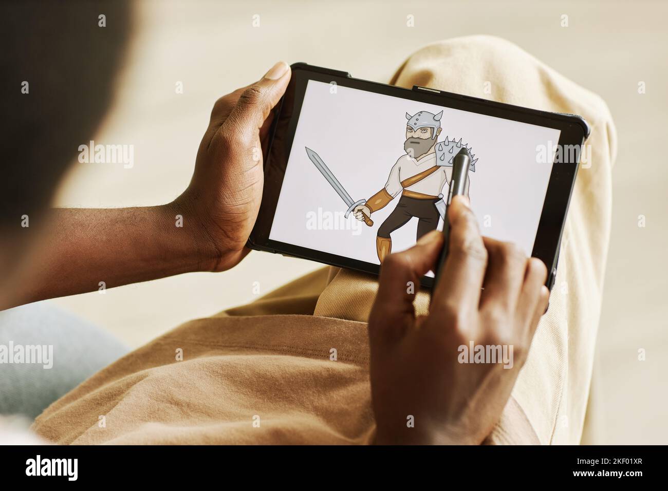Hands of young black man with stylus drawing graphic picture of warrior on tablet screen while creating new collection of digital artworks Stock Photo