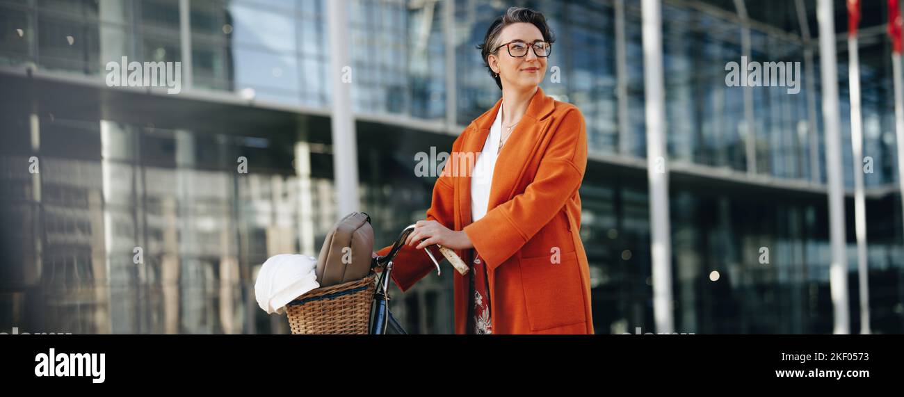 Thoughtful businesswoman pushing her bike during her morning commute in the city. Mature business woman looking away pensively while walking to work w Stock Photo