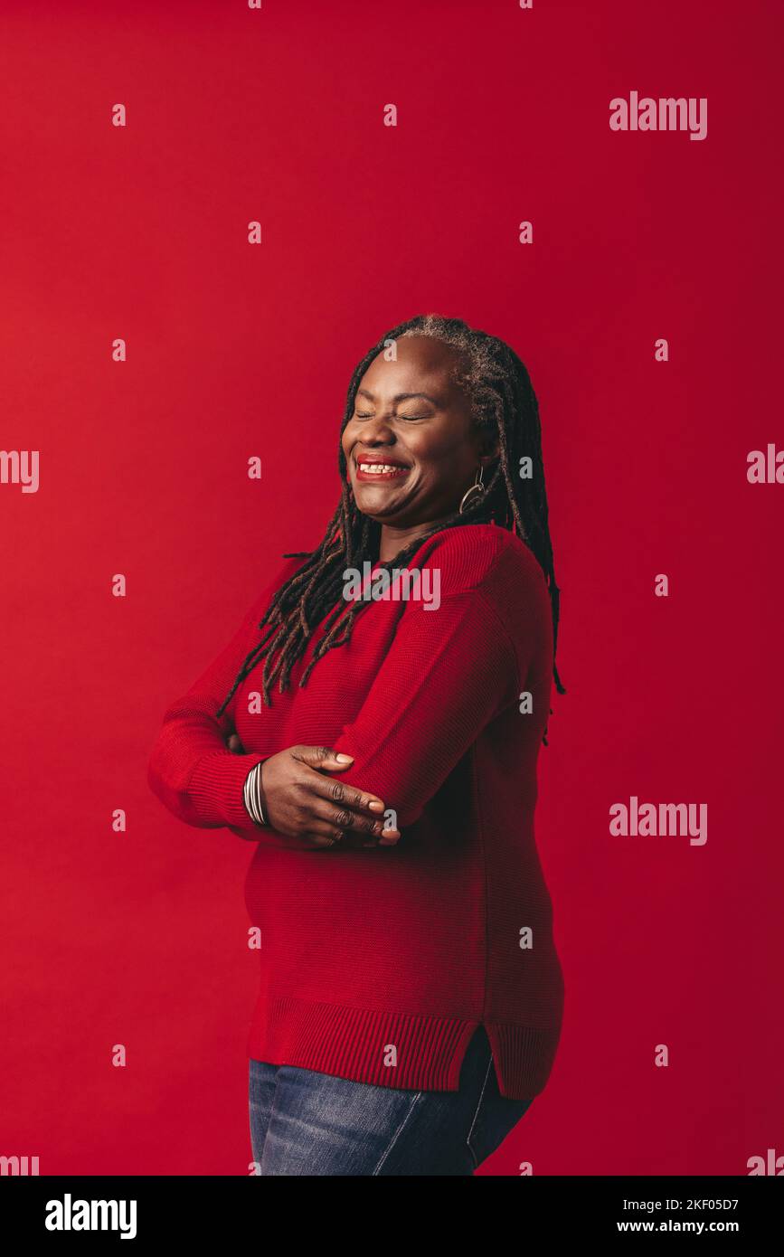 Carefree woman with dreadlocks smiling happily while standing against a red background. Mature black woman embracing her natural hair with pride. Stock Photo