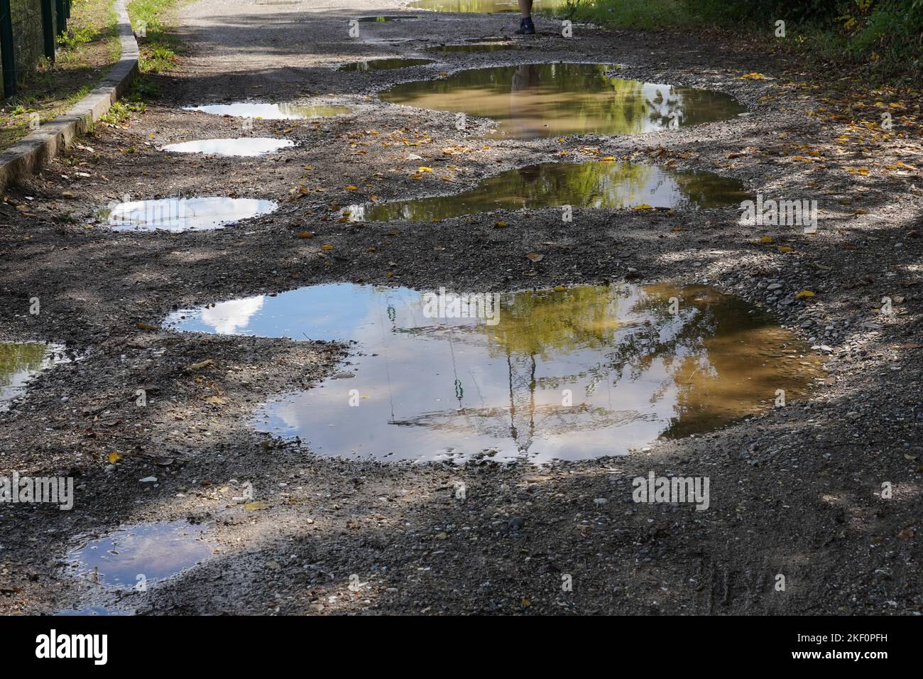 Dirt road with potholes and puddles. In the rain water of the puddles there is reflection of an electricity pillar and blue sky with some clouds. Stock Photo