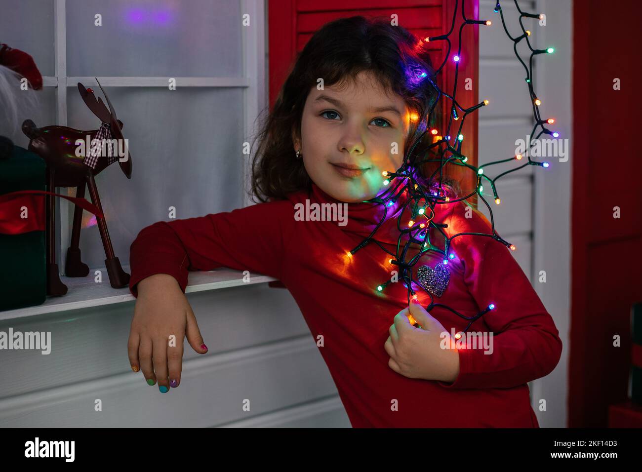 Cute little girl in red outfit hold festive colored garland leaning arm on wall. Bright lights shine in dark room. Portrait of child in decorated room Stock Photo