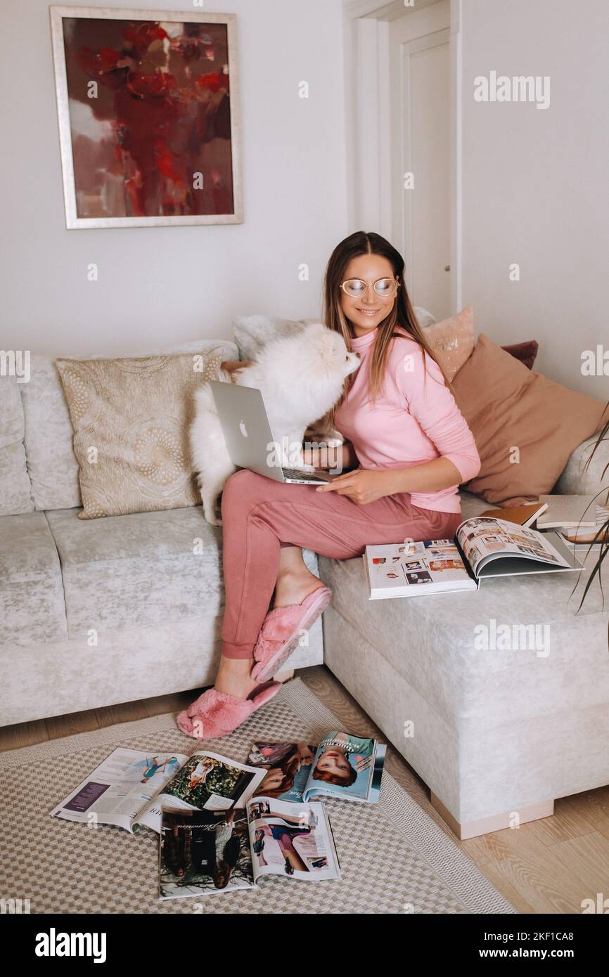 a girl in pajamas at home is working on a laptop with her dog Spitzer, the dog and its owner are resting on the couch and watching the laptop Stock Photo