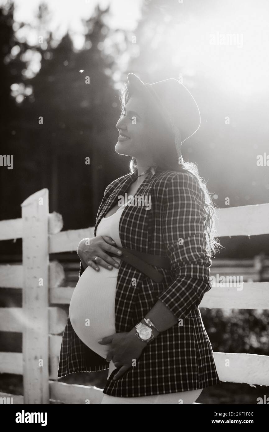 a pregnant girl with a big belly in a hat near a horse corral in nature