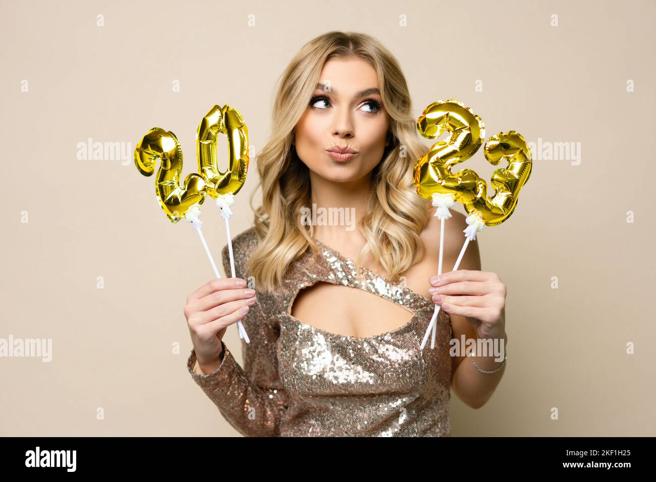 Charming young blonde girl with curly hair wearing sparkle dress holding balloons number 2023 in her hands on a beige background, new year holiday con Stock Photo