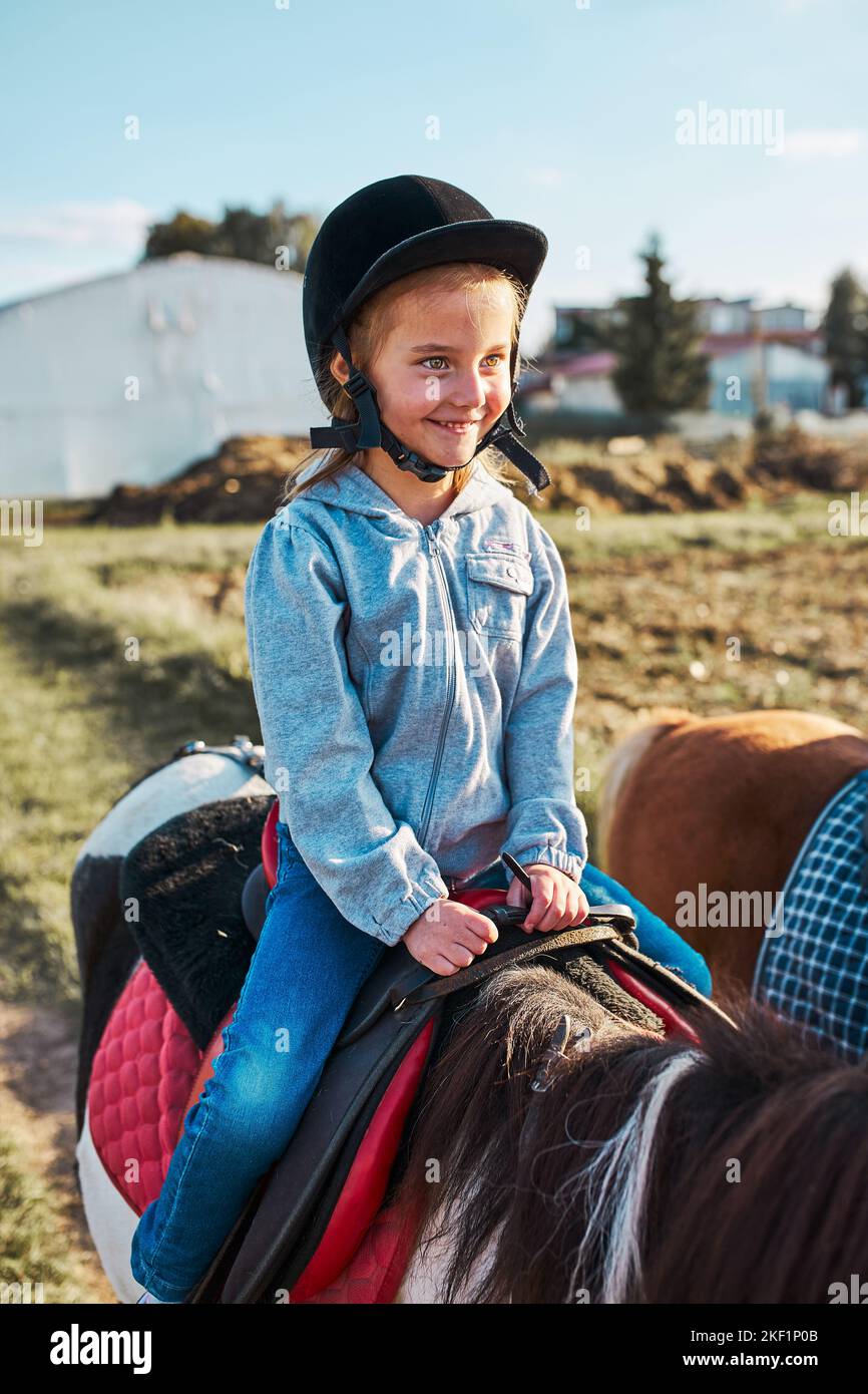 Little smiling girl learning horseback riding. 5-6 years old equestrian in helmet having fun riding a horse Stock Photo