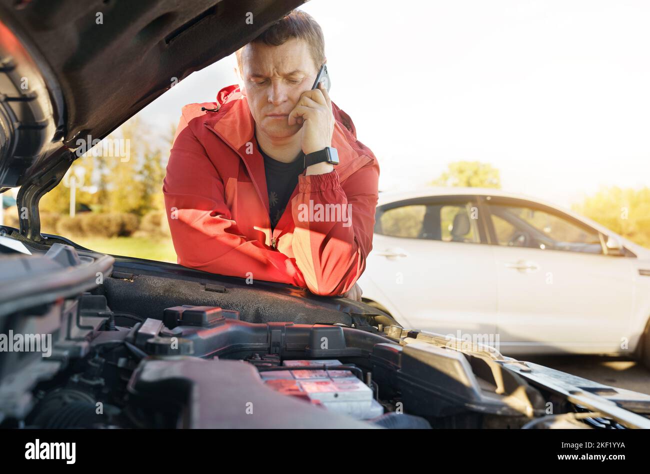 A male driver stands near the engine compartment of the car, calls the