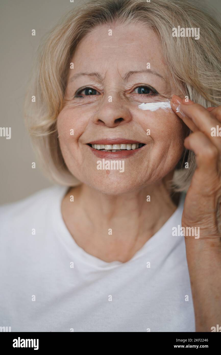 Senior woman applying moisturizing cream under her eyes on brown