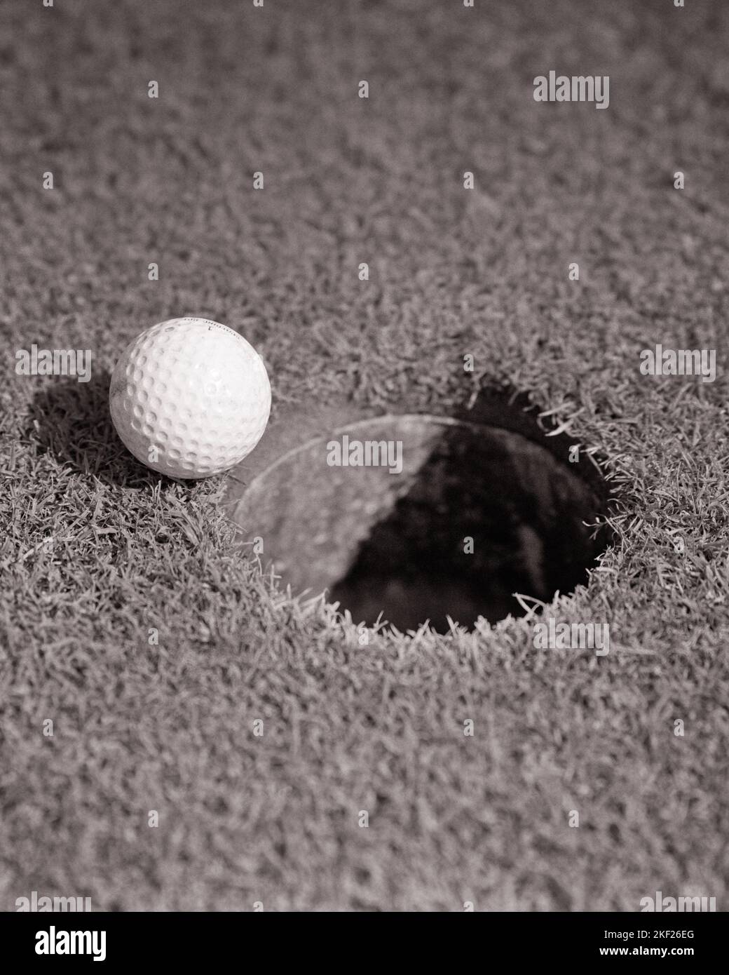 1970s DIMPLED GOLF BALL RESTING ON THE RIM OF THE CUP WITHOUT FALLING INTO THE HOLE ON THE GREEN - g8308 HAR001 HARS BIRDIE BLACK AND WHITE HAR001 OLD FASHIONED PAR REPRESENTATION Stock Photo