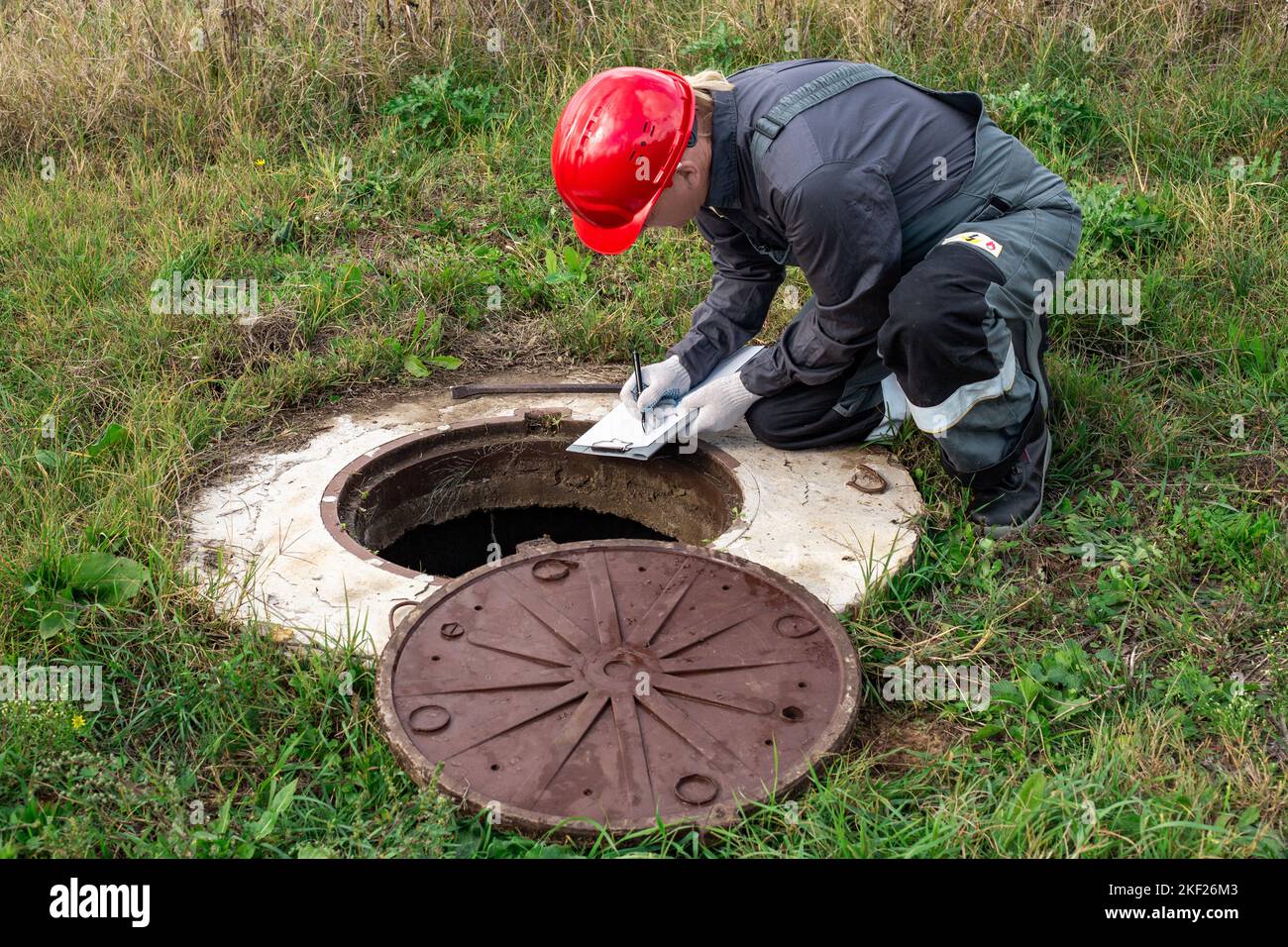 A male plumber in overalls and a helmet near a water well writes down the measurements taken and the readings of the water meter. Stock Photo
