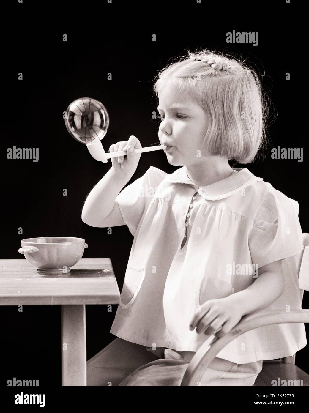 1940s LITTLE BLONDE GIRL SITTING AT TABLE BLOWING A SOAP BUBBLE FROM A