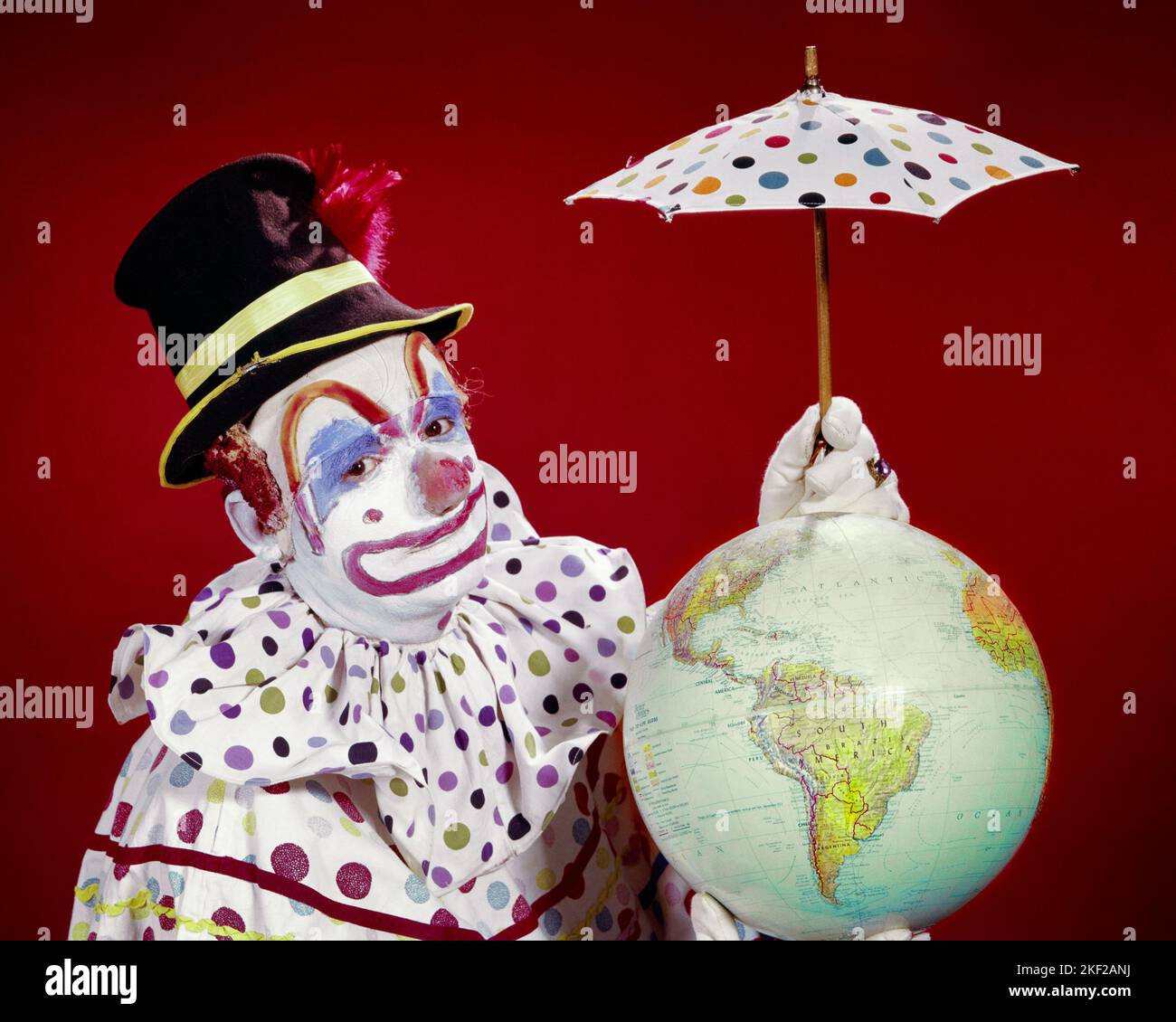 1960s CIRCUS CLOWN WEARING POLKA DOT COSTUME HOLDING A TINY PROTECTIVE UMBRELLA OVER GLOBE OF THE EARTH LOOKING AT CAMERA - kc2783 HAR001 HARS ENTERTAINMENT PROTECTIVE CLOWNS EYE CONTACT DOT GOALS PERFORMING ARTS HUMOROUS ADVENTURE ENVIRONMENT PERFORMER POLKA PROTECTION STRATEGY MAKE UP COMICAL ENTERTAINER OCCUPATIONS WHITEFACE CONCEPTUAL POLKA DOT CLOWN WHITE COMEDY JESTER SUPPORT TINY EARTH DAY TOP HAT FOOL HAR001 OLD FASHIONED Stock Photo