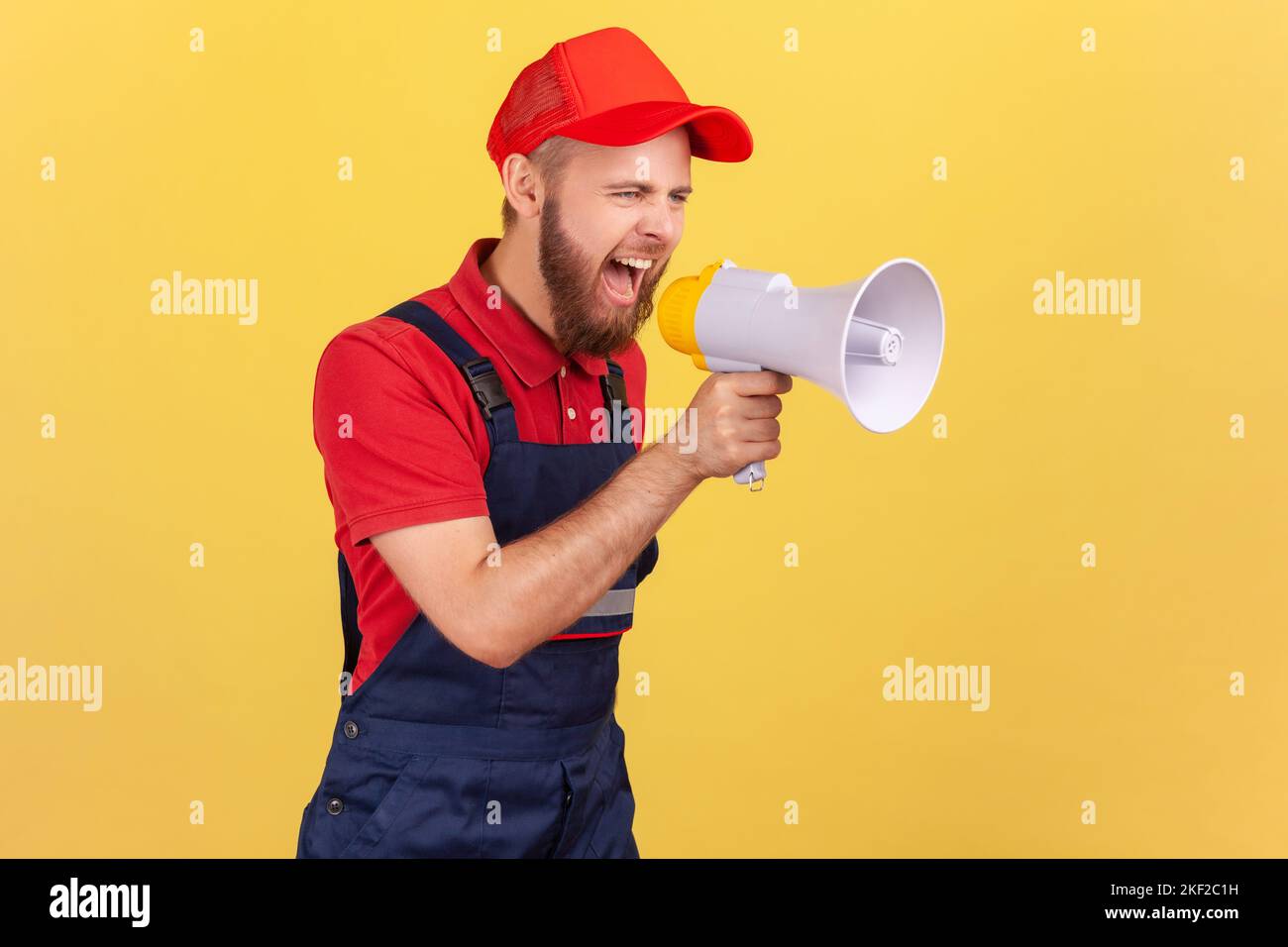Side view of angry worker man holding megaphone near mouth loudly speaking, screaming, protesting, wearing blue uniform and red cap. Indoor studio shot isolated on yellow background. Stock Photo
