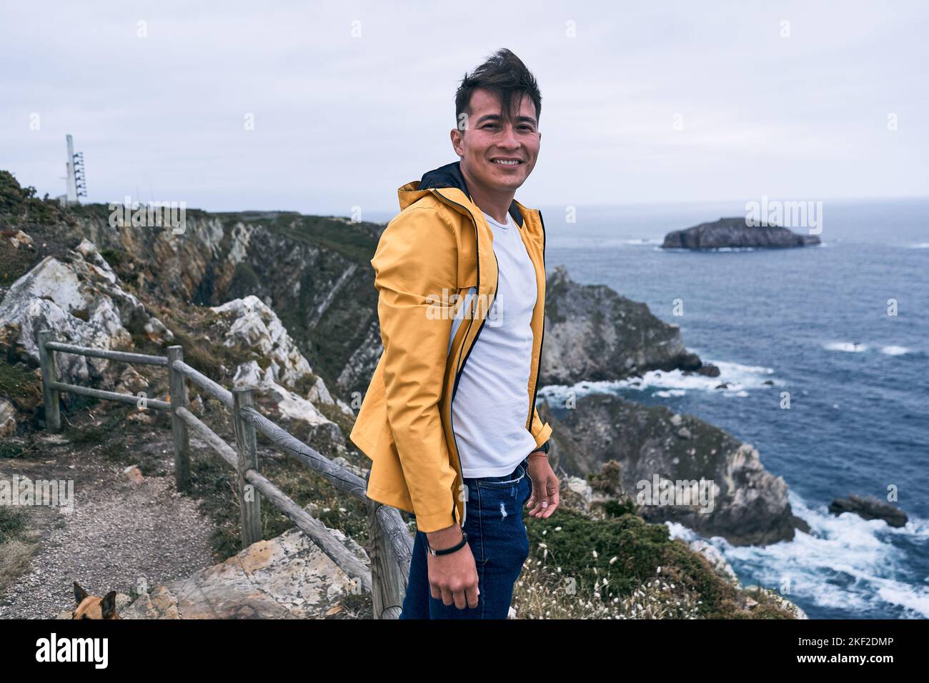 latin young man in white t-shirt and yellow jacket smiling looking at camera calm and relaxed standing in the viewpoint breathing fresh air by the sea Stock Photo