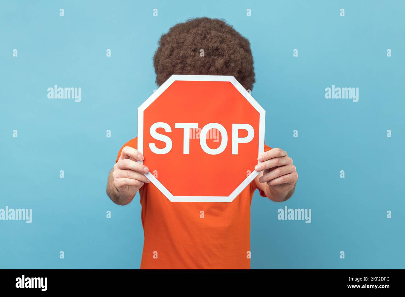 Portrait of man with Afro hairstyle wearing orange T-shirt covering face with Stop symbol, anonymous person holding red traffic sign, warning to go. Indoor studio shot isolated on blue background. Stock Photo