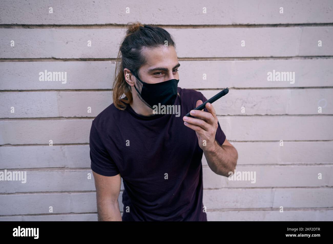 caucasian guy with long beard hair standing in black t-shirt leaning on the wall talking on the smartphone wearing face mask Stock Photo