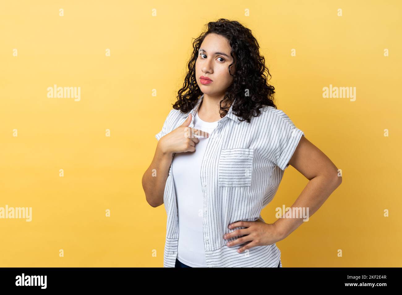 Portrait of confident woman with dark wavy hair pointing at herself, feeling proud and self-important, keeps hand on hip, having big ego. Indoor studio shot isolated on yellow background. Stock Photo