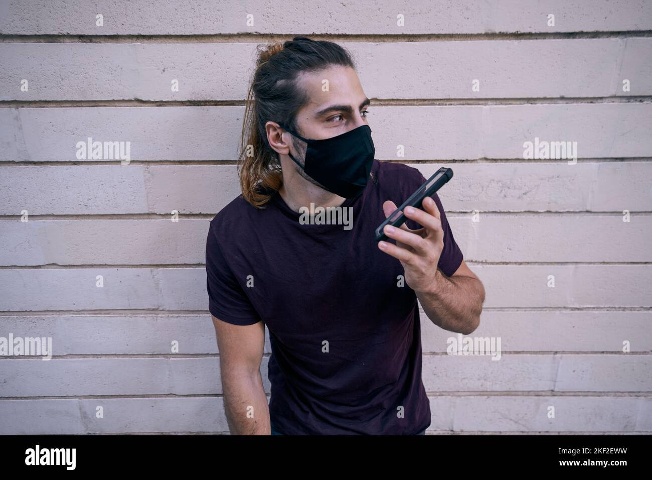 caucasian young man with long hair standing leaning against the wall talking on the smartphone wearing face mask Stock Photo